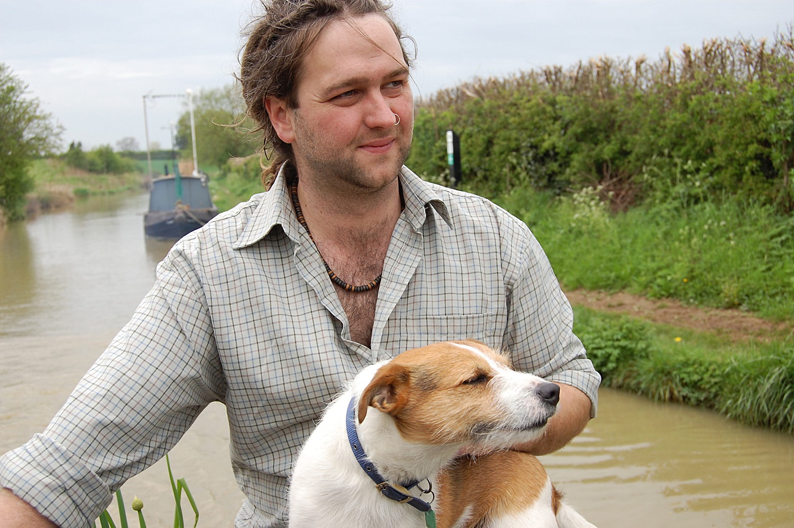 A young man sits on the back of a boat with a small dog on his lap. The dog is enjoying the breeze on his face