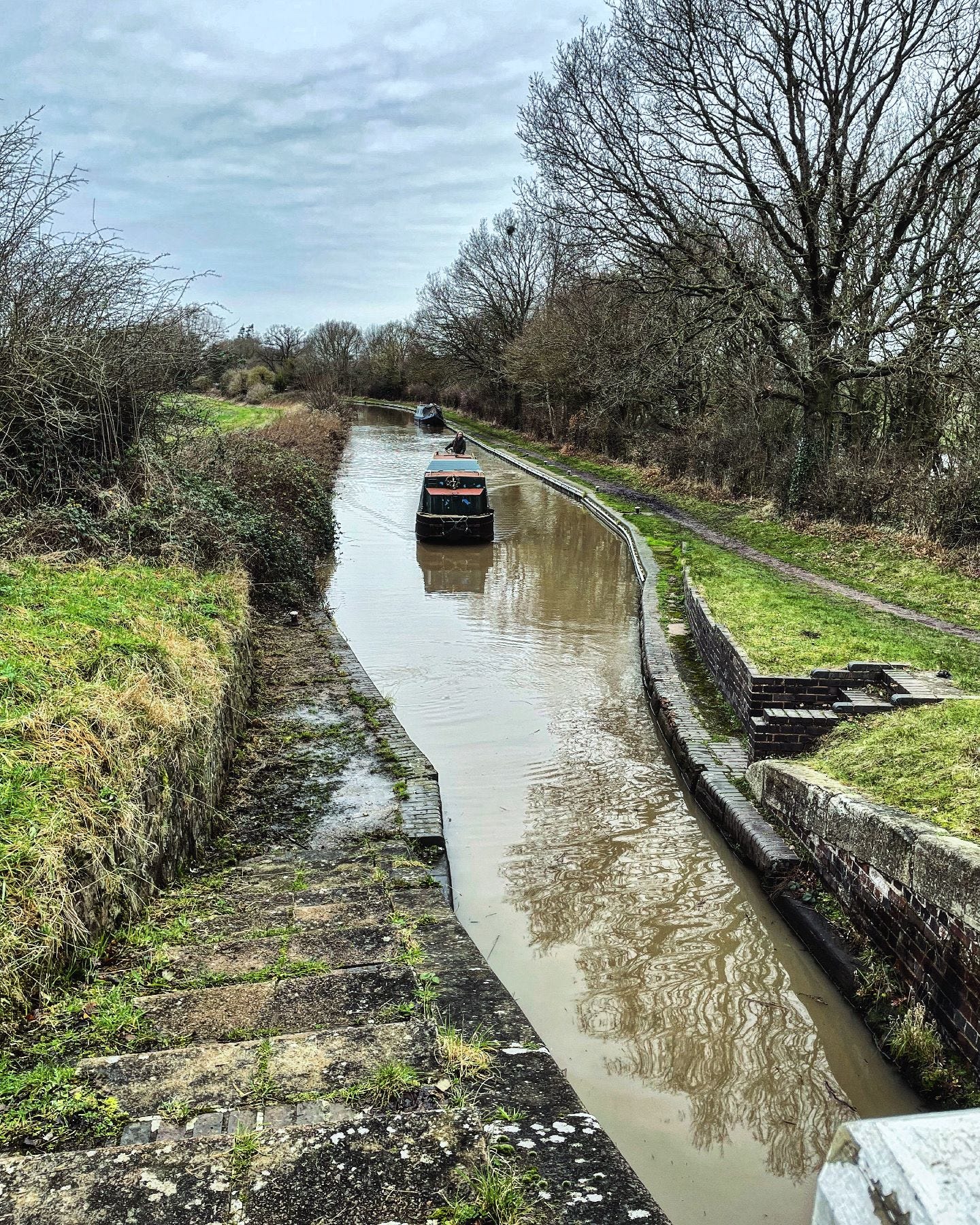 A narrowboat approaches an open canal lock. The photo is taken from above, looking down on the boat and a winter country scene of bare trees and grass