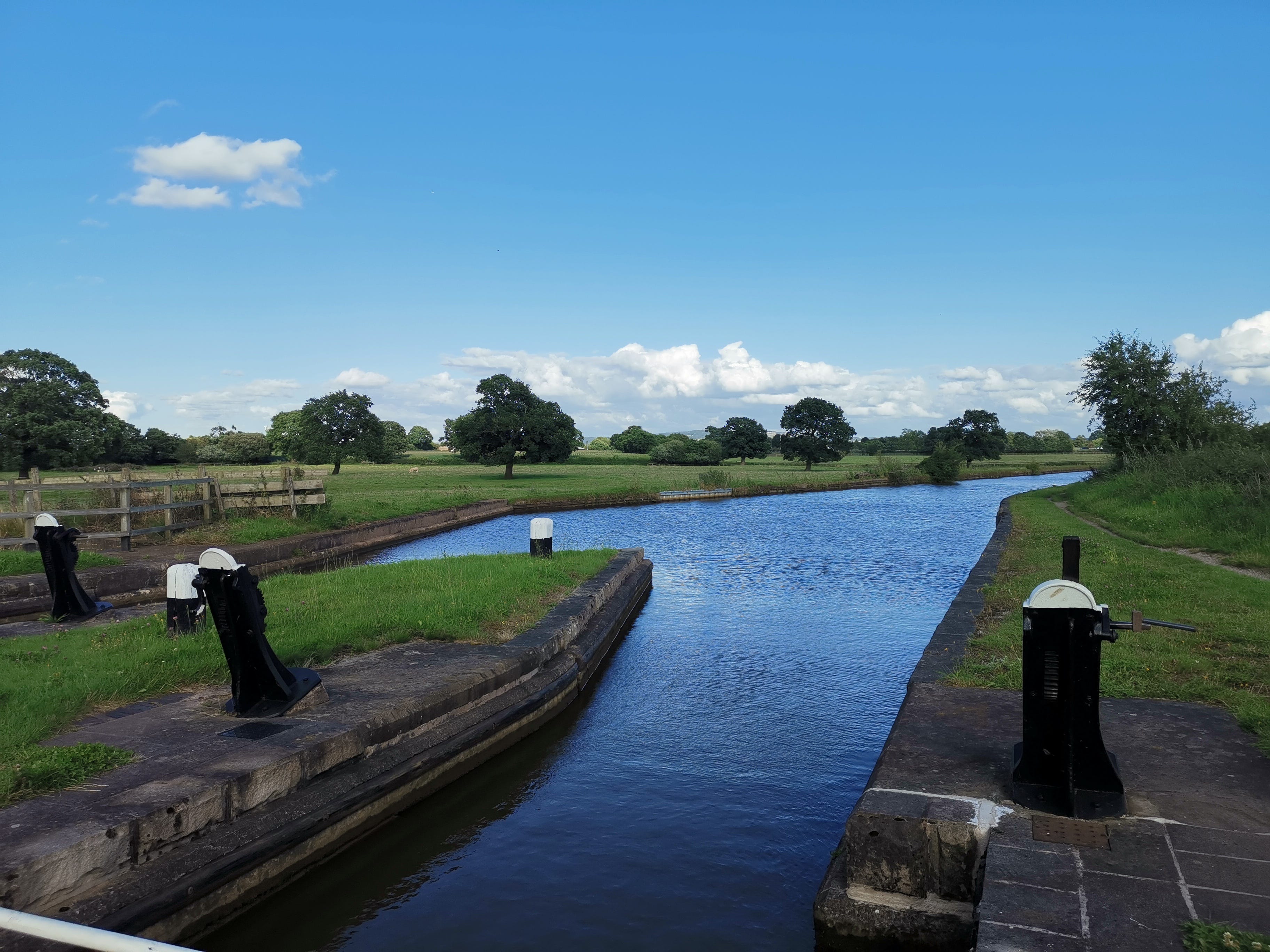 A rural view of the canal, with black and white lock gears in the foreground and green fields and trees behind