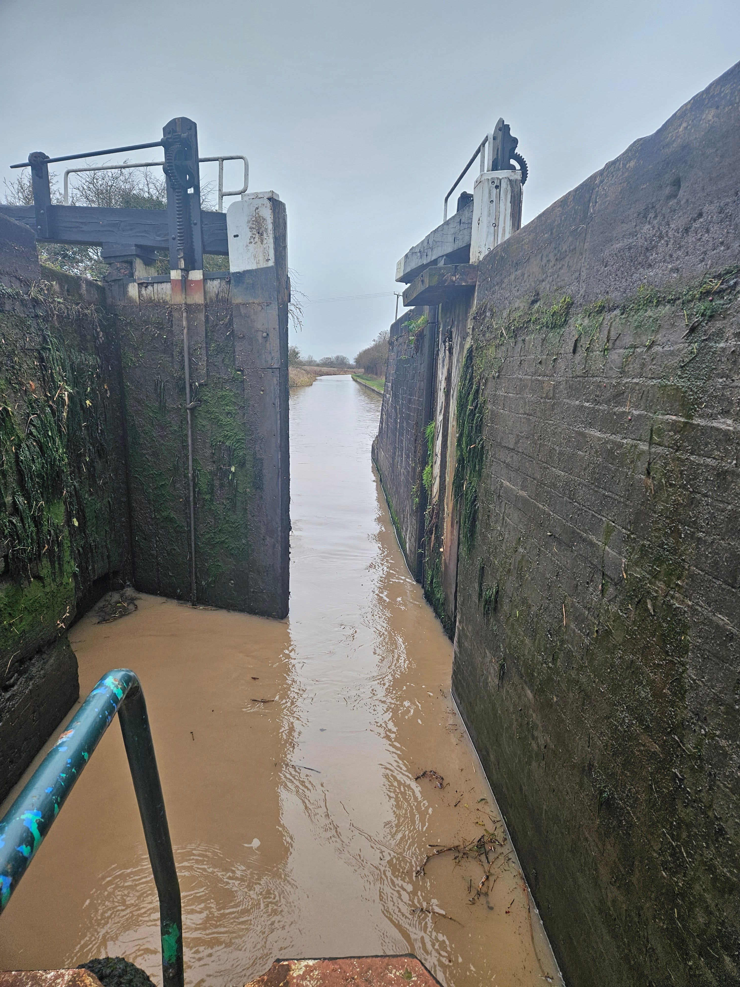 The view from the back of a boat out past open canal lock gates. Brick walls are lined with dark slime and green growth. The water is mud brown