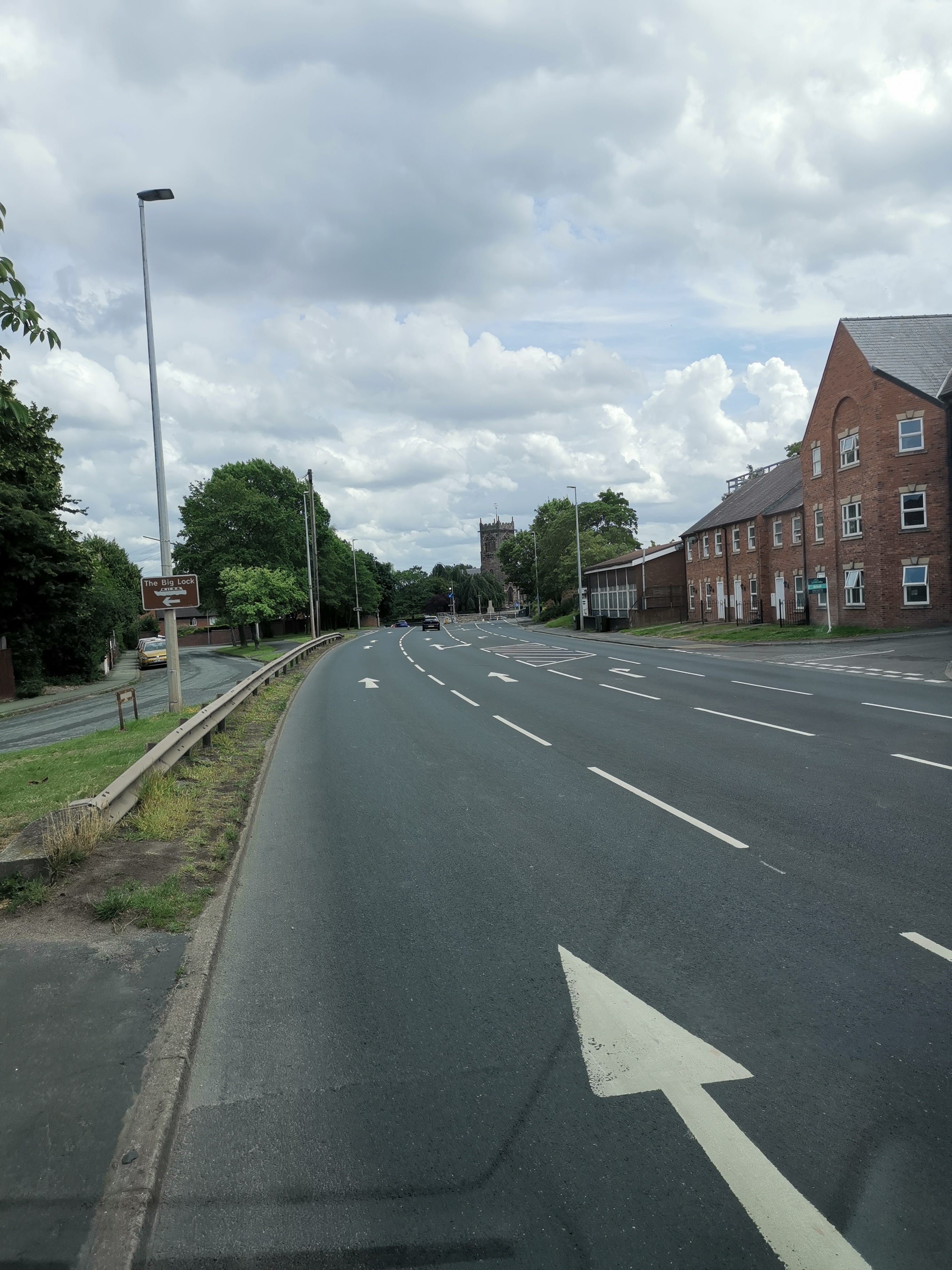 An empty four lane road leads away from the camera with town houses to the right and a side lane to the left. In the background a church spire rises from between trees in full leaf