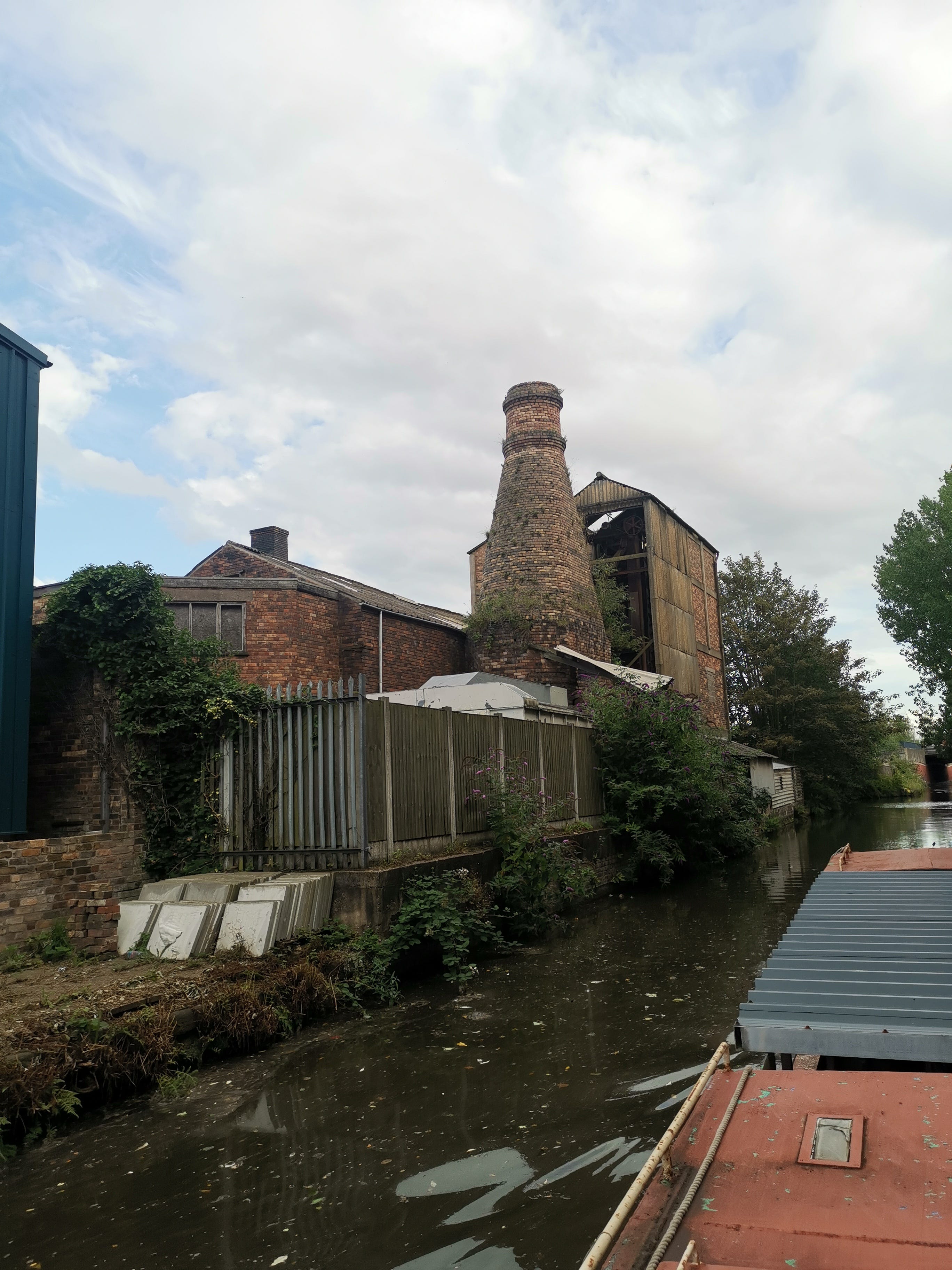 A conical brick tower with adjoining derelict factory building site alongside the canal with a small industrial unit in the foreground