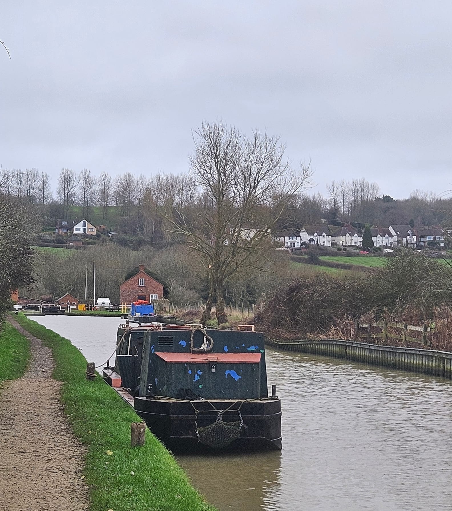 A Black and green narrowboat, a working boat with steel cabin, lies tied up against a grassy towpath. Behind it is a line of white houses on a rural hillside, the image is crowned with trees at the top of the hill