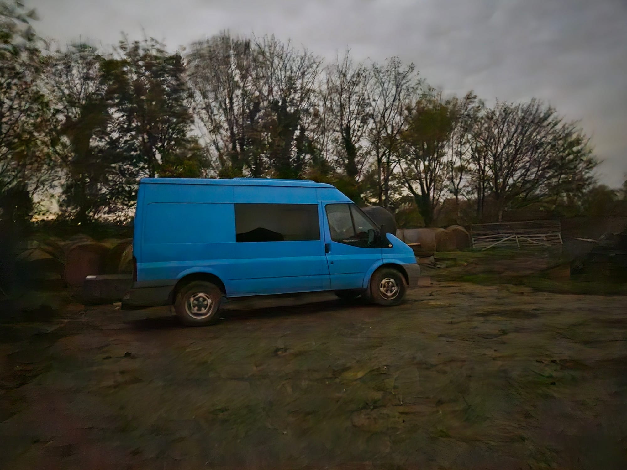 a blue van with black windows is parked in dim light at the edge of a farm yard, with trees behind